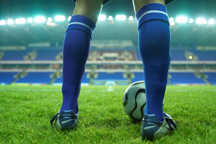 A close-up of a person standing on a grass field with a soccer ball at their feet, viewed from behind, in an empty soccer stadium