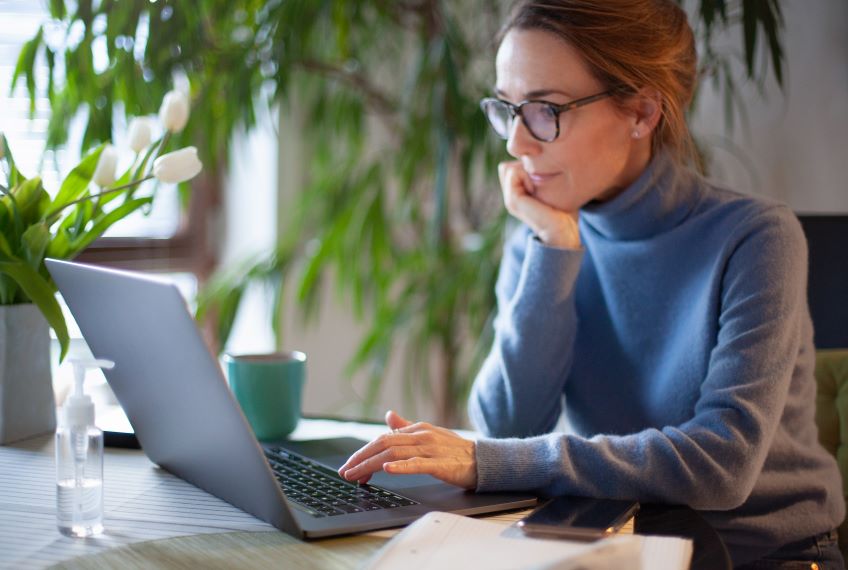 An individual sits at a table using a laptop, with a cup, a bottle of hand sanitizer and some papers beside the laptop