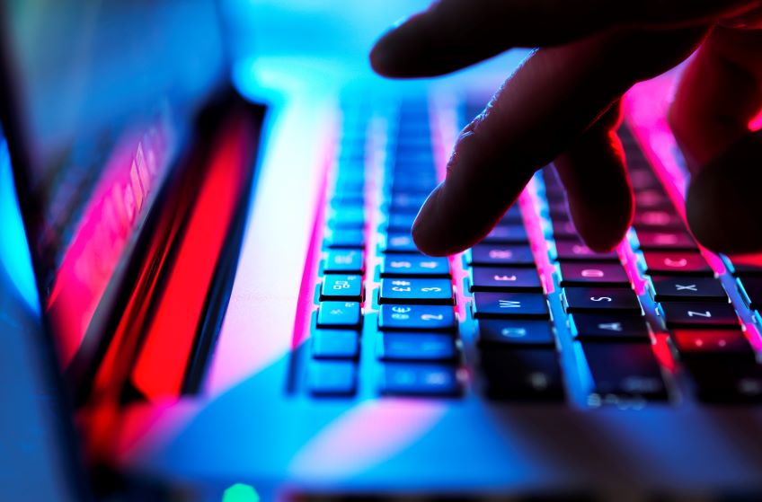 A close-up of a person’s hand typing on a laptop keyboard, which is backlit with blue and red colors