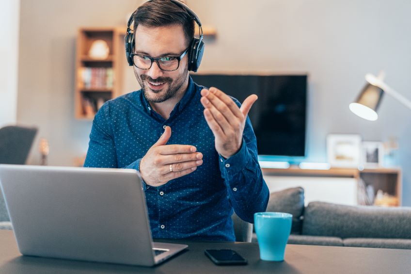 An individual sitting at a table with a laptop open in front of them, wearing headphones and gesturing with their right hand, with a blue cup and a smartphone on the table