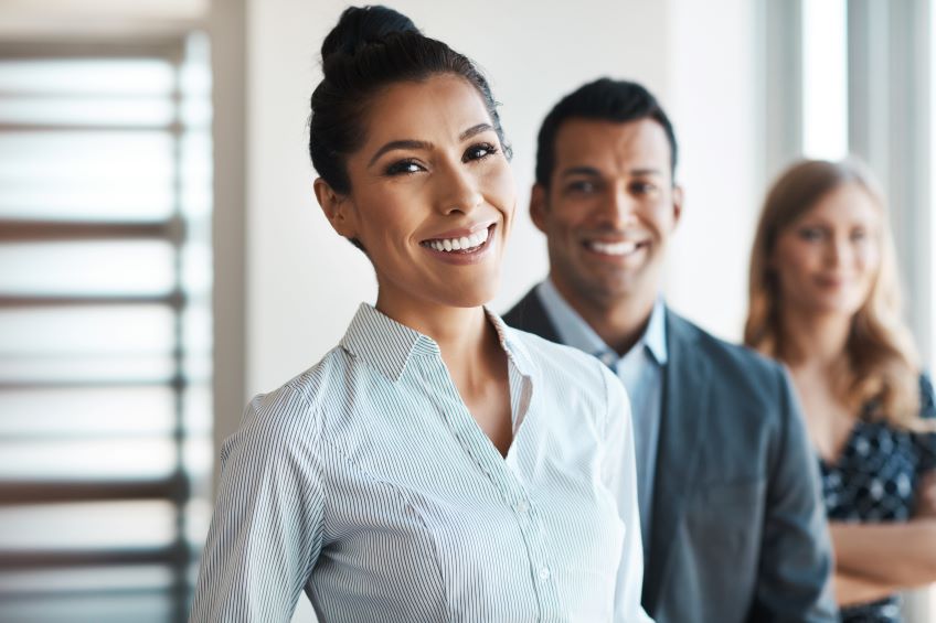 Three individuals are standing in a professional environment, smiling into the camera and staggered in position