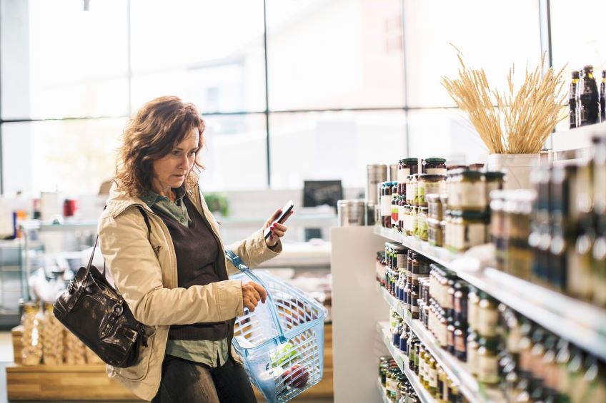 A person standing in a grocery store aisle holding a smartphone and a shopping basket filled with various items