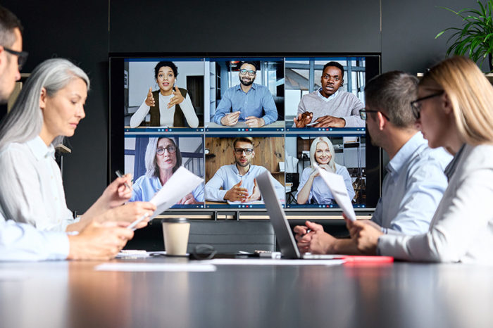A group of four people seated at a table having a video conference call with six people who are shown on a large monitor.