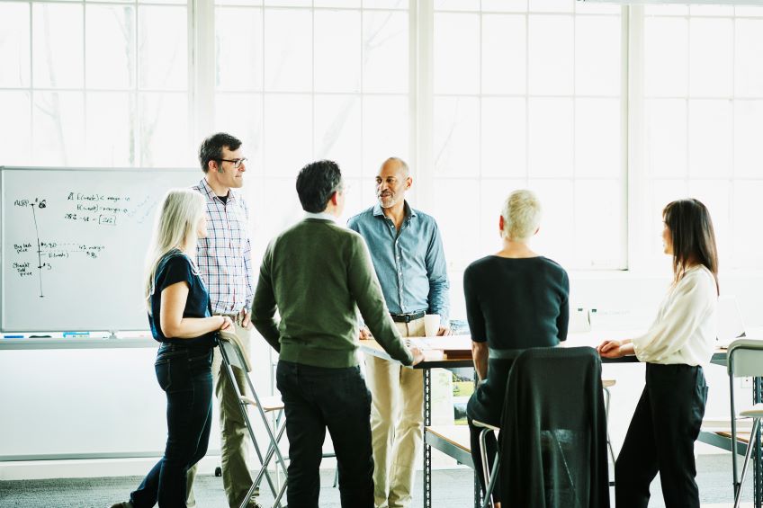 A group of co-workers in discussion stands in front of a whiteboard.