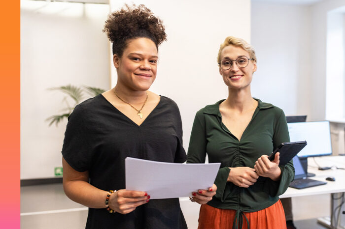 Two women in casual business attire smile at the camera in an office setting