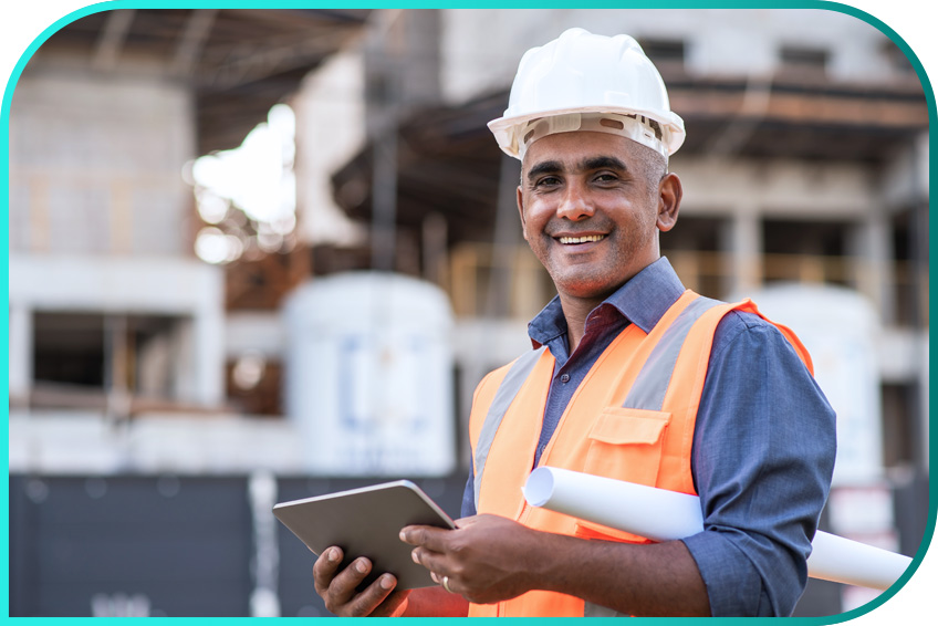 A construction worker with a safety helmet and orange vest is holding a tablet and rolled-up blueprints at a construction site