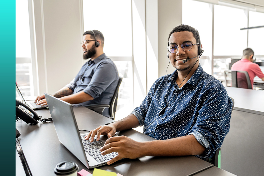 Two individuals wearing headsets work on laptops in a call center setting as one looks at the camera and smiles and a third person works at a desk in the background