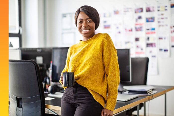 A person holding a mug leans against a table with computers and smiles