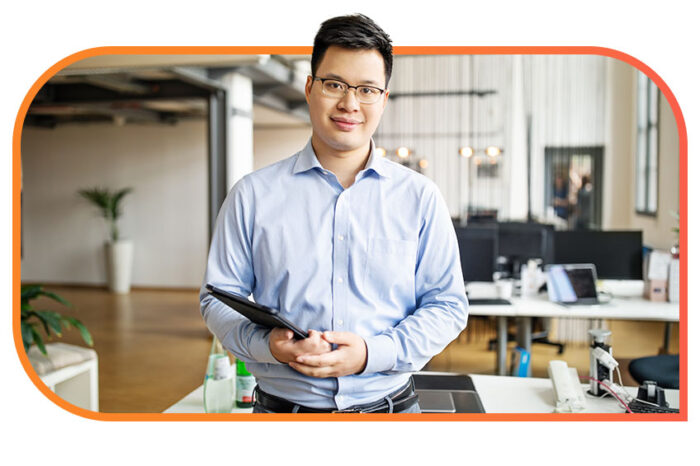 A person in business casual attire holds a tablet device in an office and looks at the camera