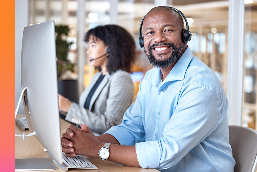 Two individuals in an office setting working on desktop computers, one smiling and facing the camera while the other looks at their computer screen