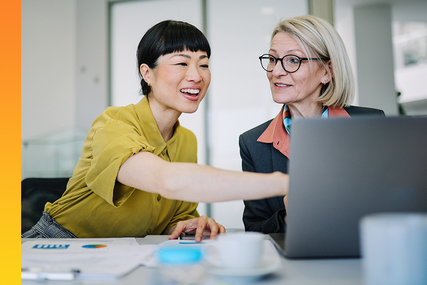 Two office employees smile and discuss business in front of laptops on a table