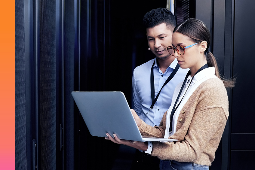 Two employees in business attire look at a laptop in a server room